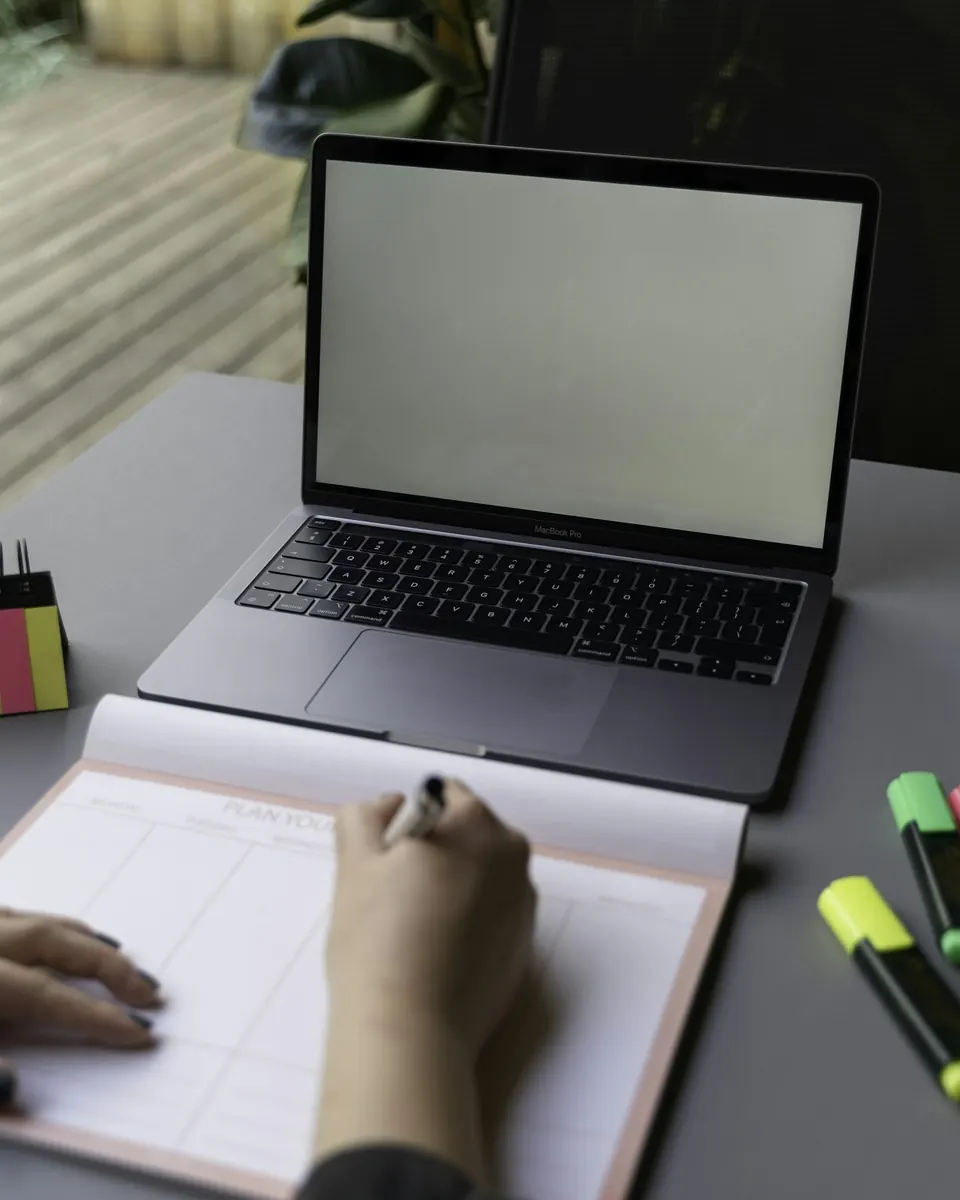 Person plant ein Webdesign sorgfältig mit Stift und Papier neben einem Laptop